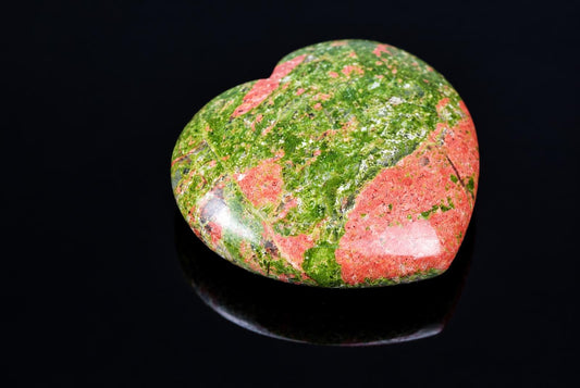 A polished heart-shaped stone with green and pink patterns on a dark reflective surface.