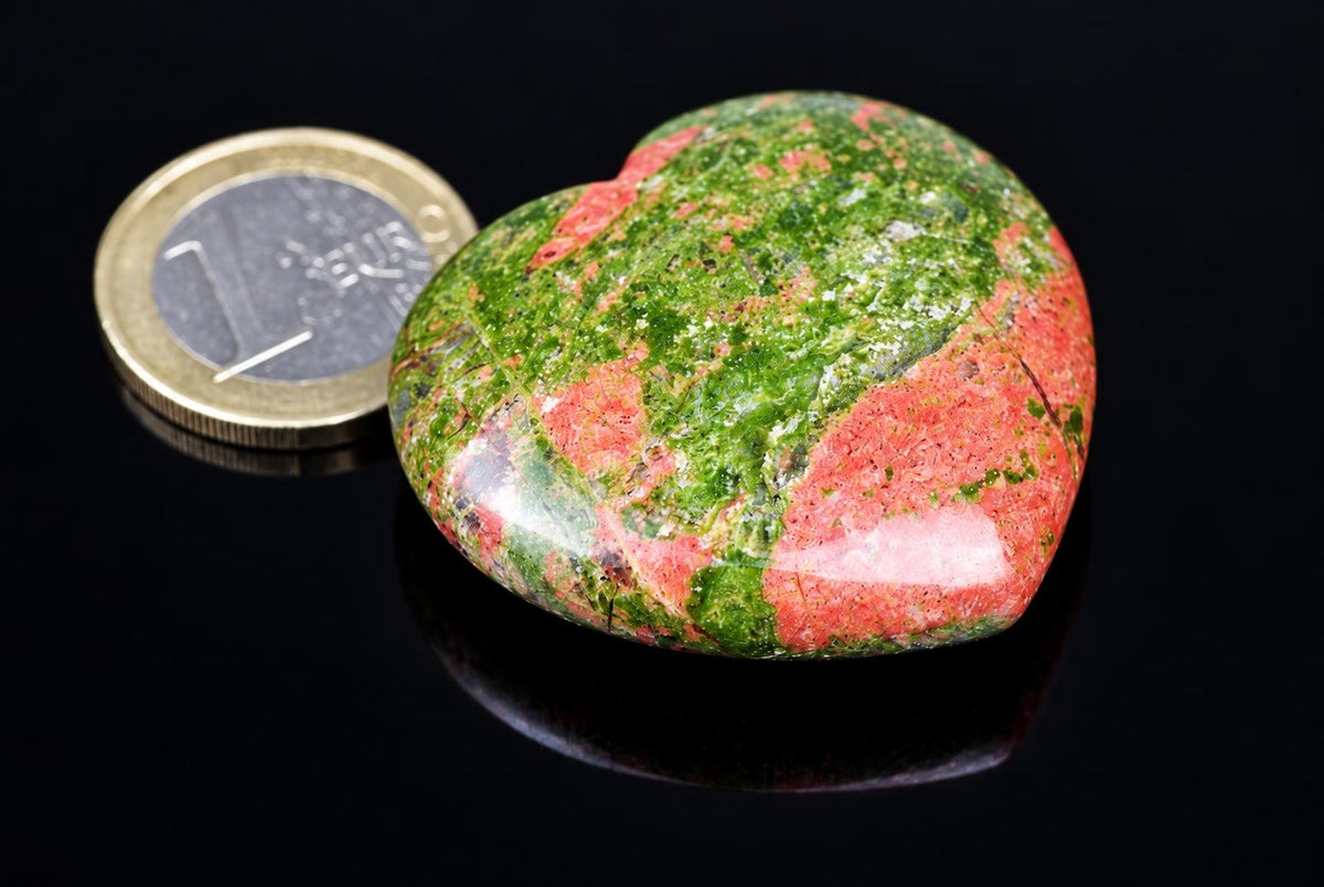 A polished heart-shaped stone with green and red patterns sits on a dark surface next to a coin.