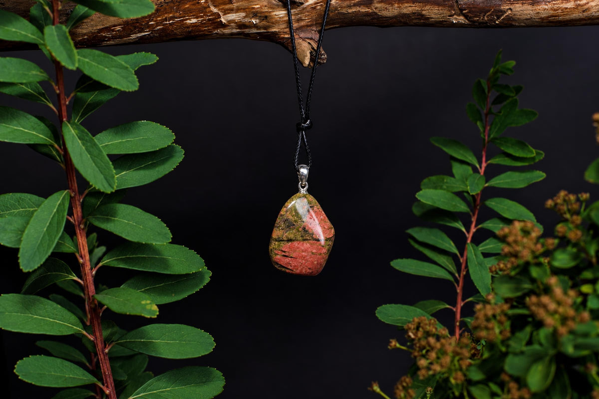 A polished unakite pendant with pink and green stripes hangs from a black cord on a wooden branch, framed by green leaves.