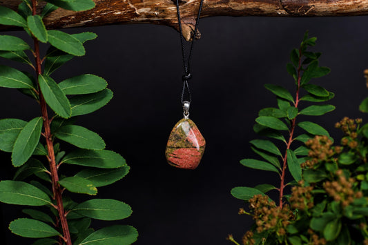 A polished unakite pendant with pink and green stripes hangs from a black cord on a wooden branch, framed by green leaves.