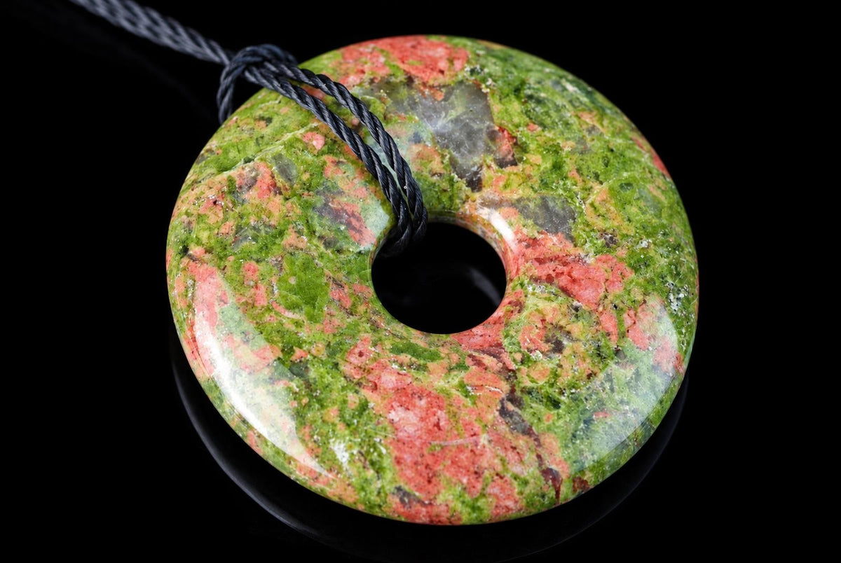 A polished donut-shaped pendant with green and pink speckled stone, a central hole, and a black cord looped through it on a dark surface.