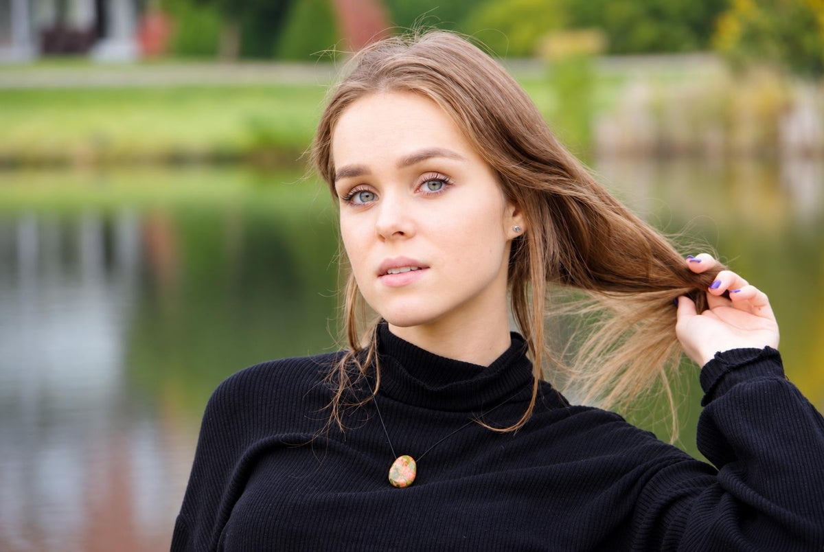 A woman wears a black turtleneck sweater with an oval pendant featuring green, pink and brown patterns on a thin cord.