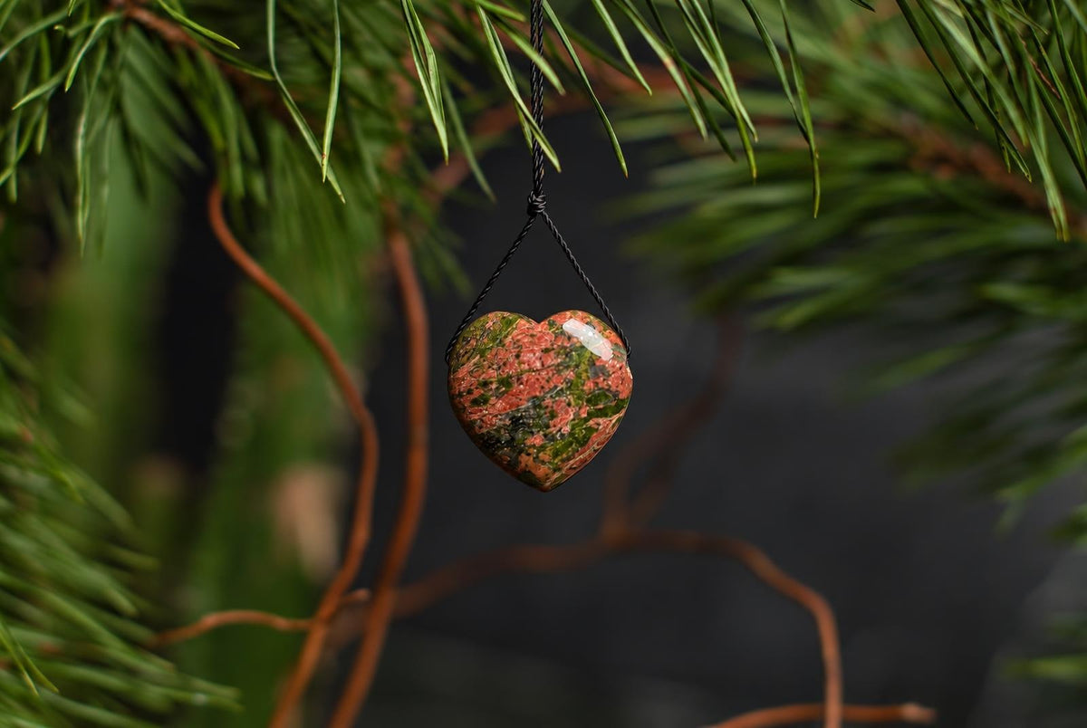 A heart-shaped pendant with orange and green mottled patterns hangs from a black cord among green foliage and dried stems.