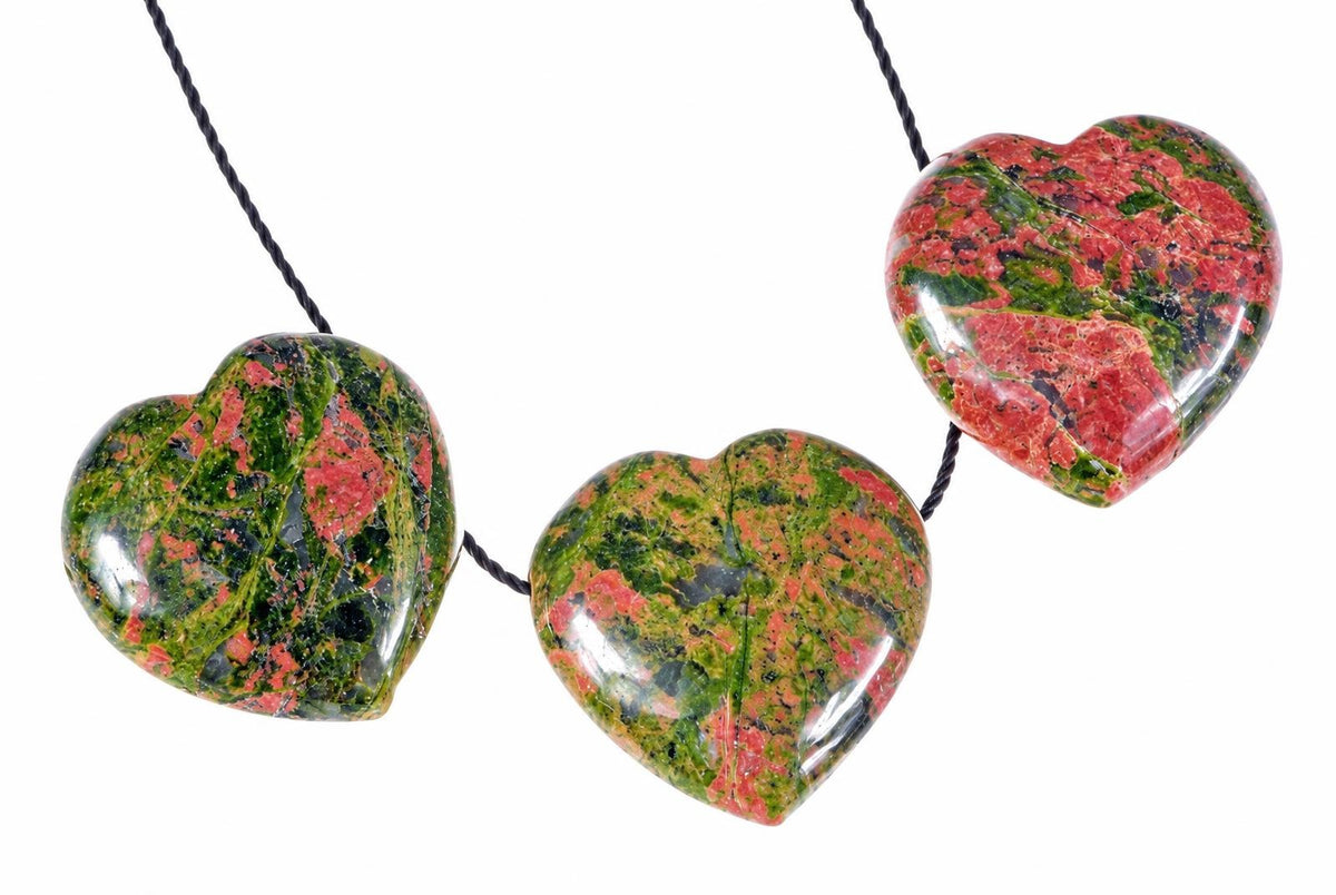 Three polished heart-shaped unakite pendants with red, green and black patterns hang on a dark cord against a white background.
