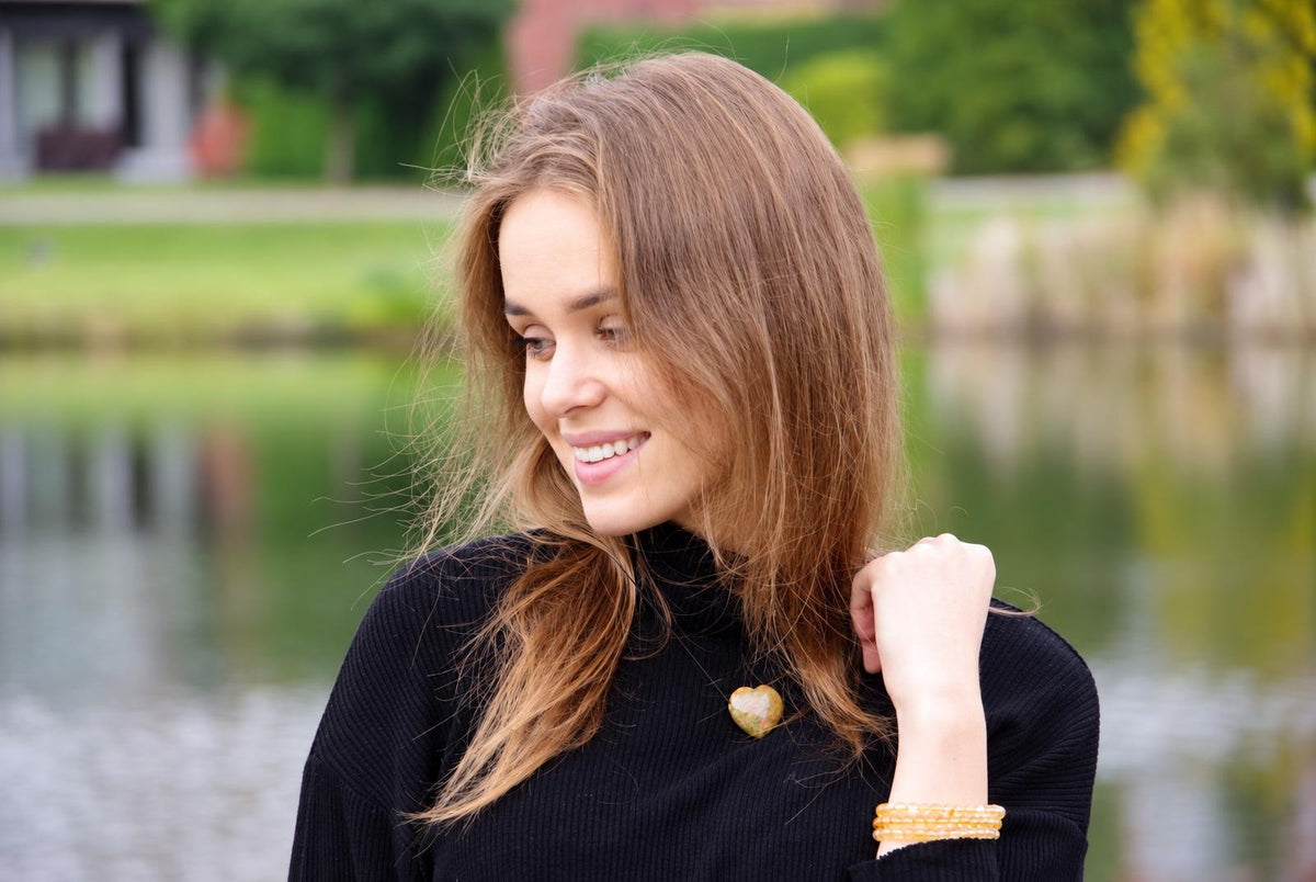 A woman wears a black turtleneck with a heart-shaped unakite pendant and yellow beaded bracelets, outdoors near water.