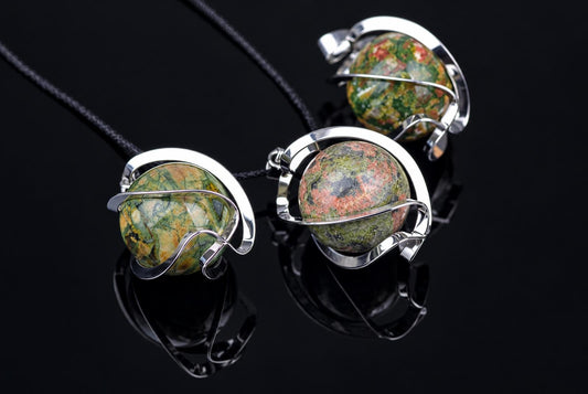 Three pendants with round, multicoloured unakite spheres in silver-toned wire settings on dark cords against a black reflective surface.