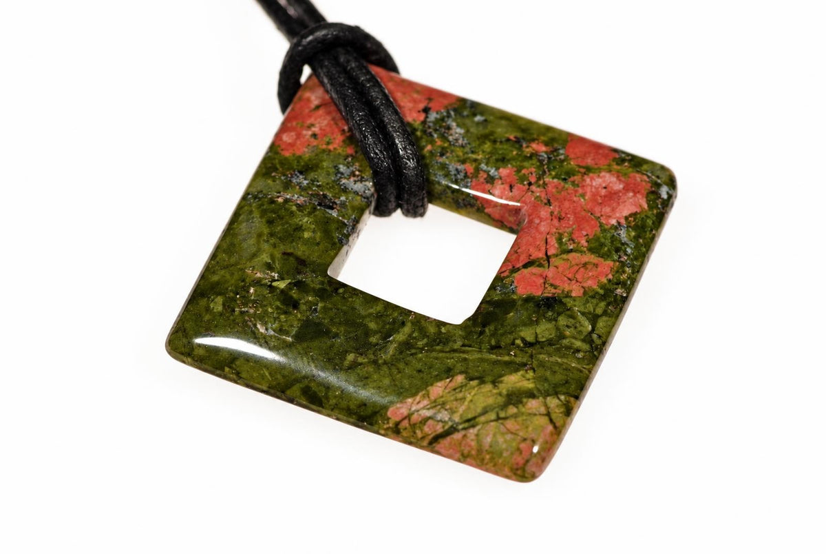 A square unakite pendant with a central hole, featuring green and red patterns, on a black cord against a white background.