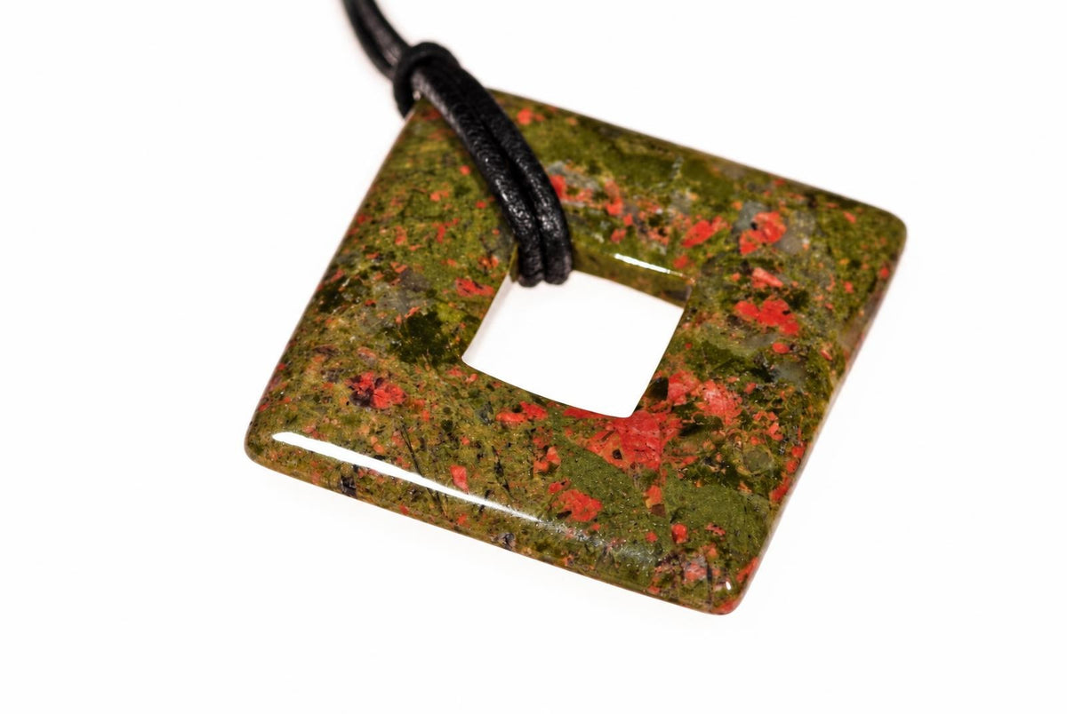 A square unakite pendant with a central hole, featuring green and red speckles, on a black cord against a white background.