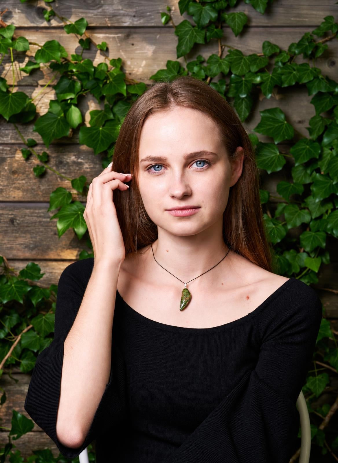 A woman wears a black top and a pendant with a green, wing-shaped stone on a dark cord against a wooden wall with ivy.