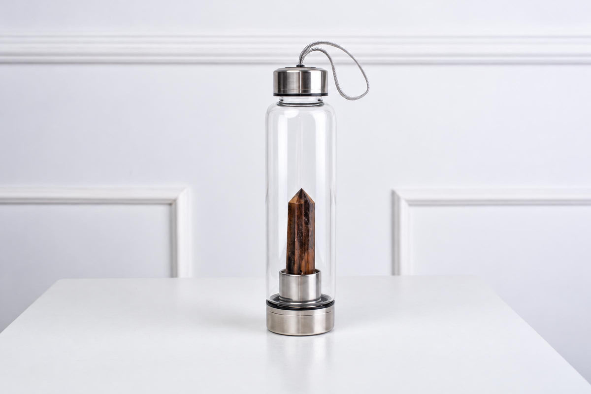 A clear glass water bottle with a silver-toned lid and base, containing a brown obelisk crystal inside, on a white surface.