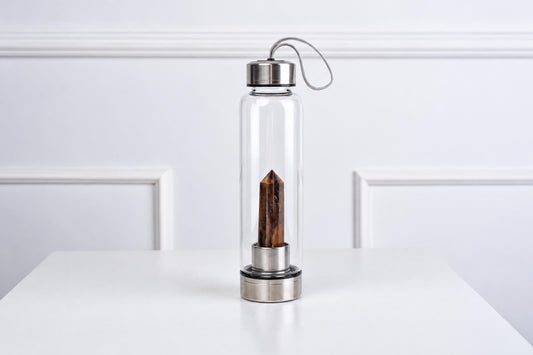 A clear glass water bottle with a silver-toned lid and base, containing a brown obelisk crystal inside, on a white surface.