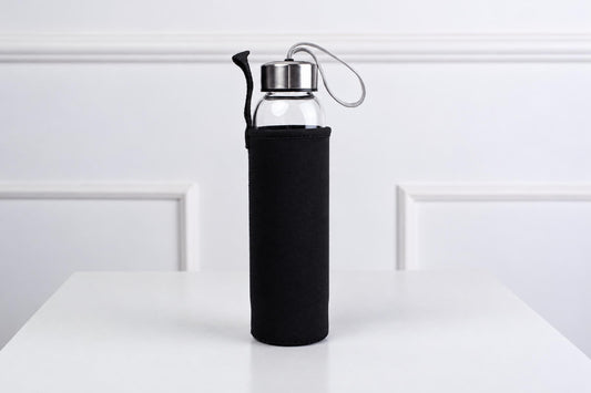 A glass water bottle with a silver-toned lid and black fabric sleeve stands on a white surface.