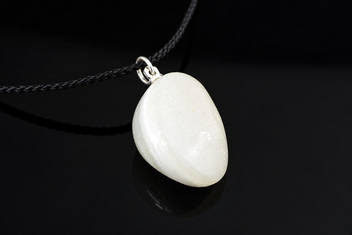 A white oval pendant with a silver-toned bail hangs from a black cord on a dark reflective surface.