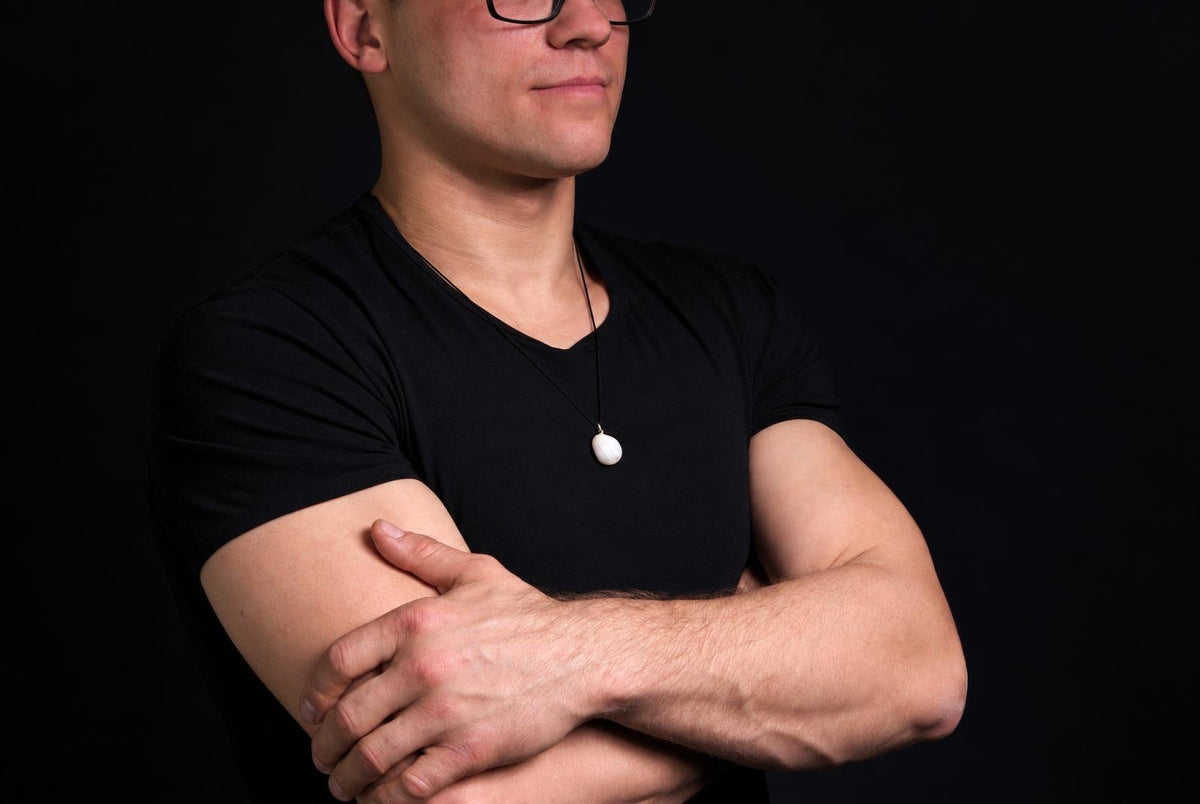 A man in a black t-shirt wears a white oval pendant on a dark cord against a black background.
