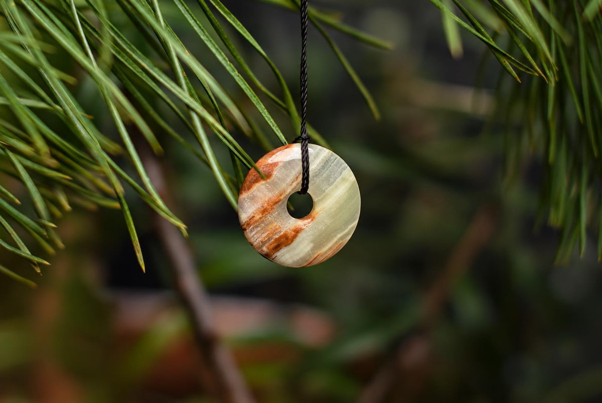 A pale green and cream circular stone pendant with a central hole and orange-brown bands hangs from a black cord among green foliage.