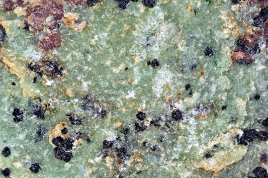 A close-up of a green zoisite raw stone with black, white, and brown mineral inclusions on a dark surface.